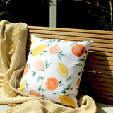 Les Fruits Abstract Polyester Cushion in multi, a 43 x 43cm square cushion with watercolour lemons, oranges and stone fruits among leaves on white, shown on an outdoor wooden bench with a woven throw.
