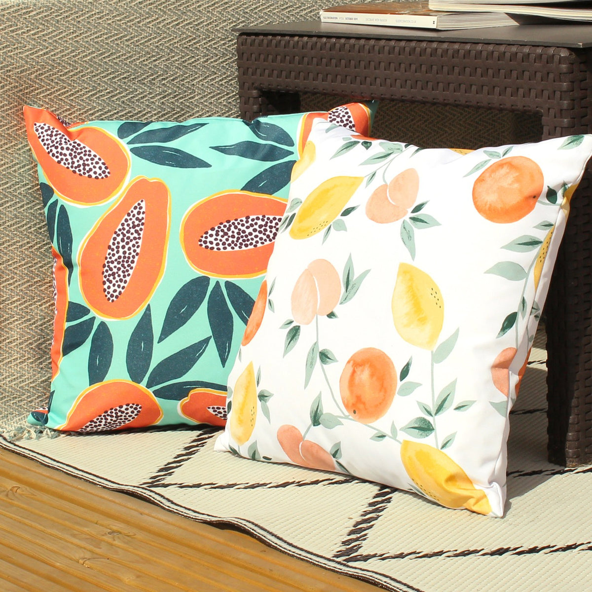 Les Fruits Abstract Polyester Cushion in white with lemons, oranges and stone fruit design, shown beside a papaya-print cushion on an outdoor rattan seating area.