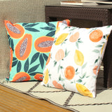 Les Fruits Abstract Polyester Cushion in white with lemons, oranges and stone fruit design, shown beside a papaya-print cushion on an outdoor rattan seating area.