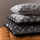 Stack of three Lexington jacquard velvet cushions in grey and black geometric patterns—diamond, hexagon, and chevron designs—displayed on an orange tufted ottoman.