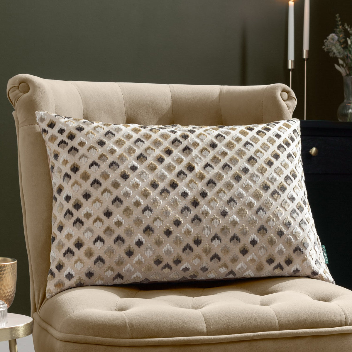 Lexington Jacquard Velvet Cushion in Warm Taupe with a diamond-patterned gold, charcoal and grey design, styled on a beige tufted armchair.