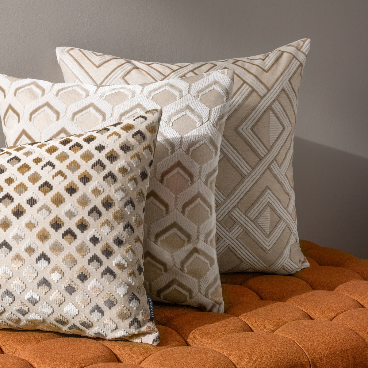Lexington Jacquard Velvet Cushion in warm taupe with a diamond-patterned rectangular design, shown alongside geometric-print cushions on an orange tufted seat.
