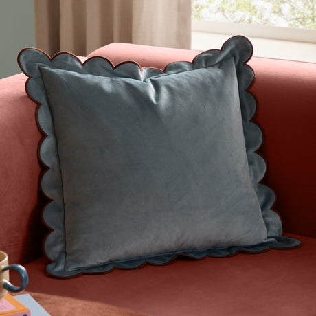 Lil Scallop Plain Velvet Cushion in blue with scalloped edge and contrasting brown trim, shown on a pink sofa in a living room setting.