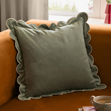 Lil Scallop Plain Velvet Cushion in sage green with a scalloped edge and contrasting natural stitching, shown on an orange sofa beside a curtained window.