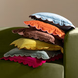 Lil Scallop velvet cushion in orange with pink scalloped edge trim, shown stacked with other scallop-edge cushions on a green sofa.