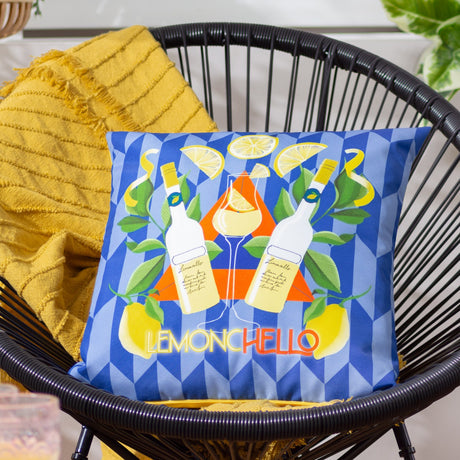 Limoncello Abstract Polyester Cushion in blue, printed with lemons, leaves and Limoncello bottles on a geometric diamond ground, shown on a black wicker chair with a mustard throw.