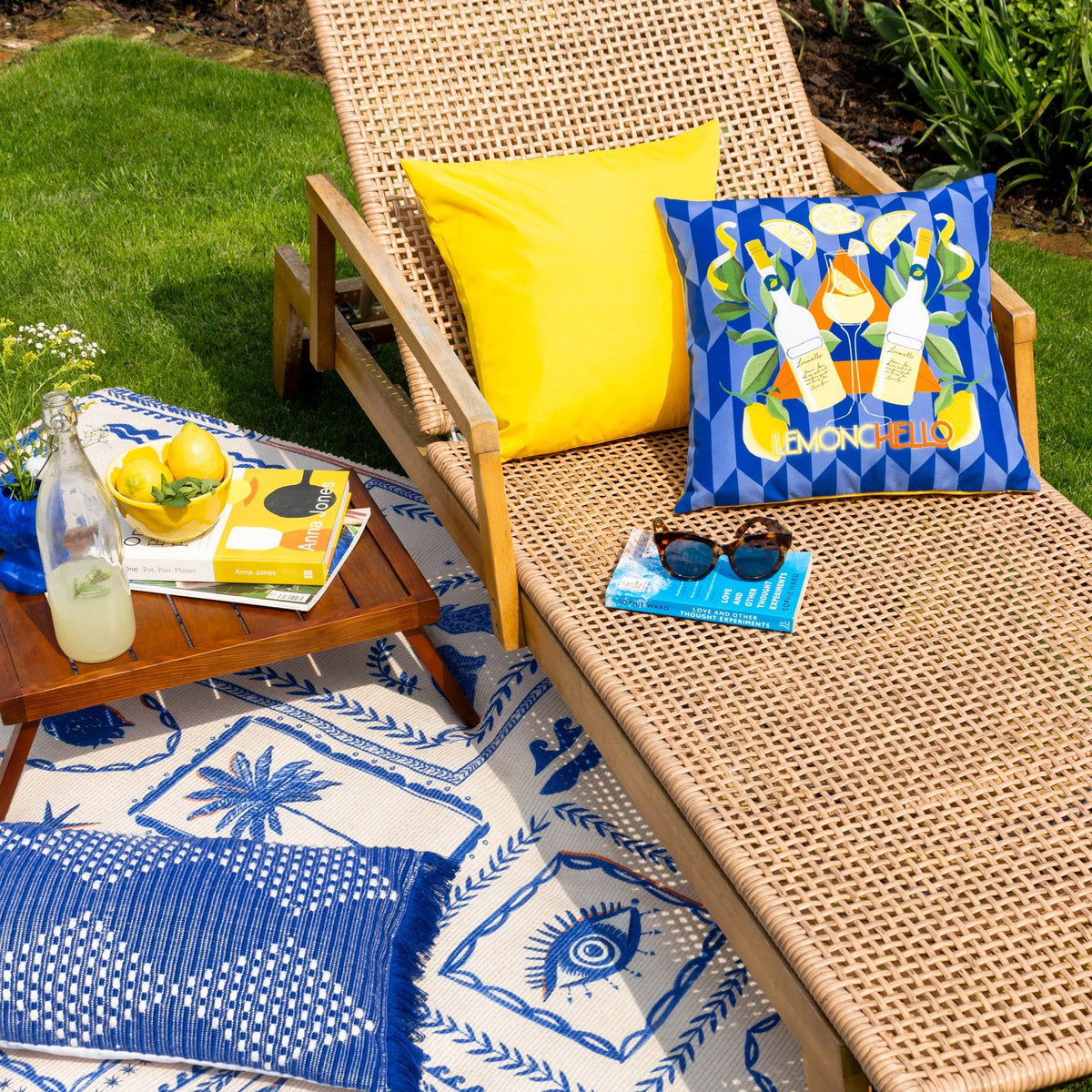 Limoncello Abstract Polyester Cushion in blue with orange reverse, featuring lemon and bottle motifs, styled on a wicker garden lounger outdoors.
