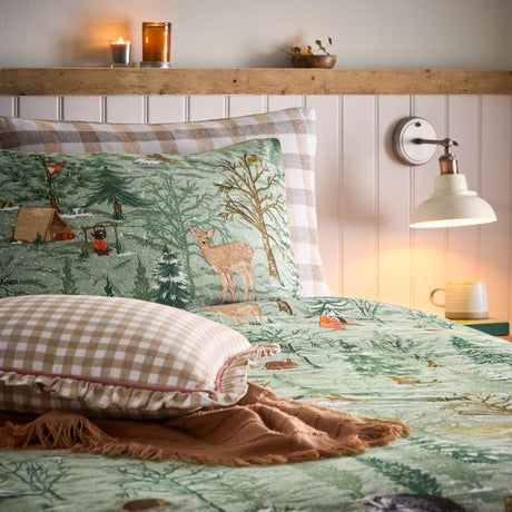 Lodge Wood Cotton Animal Duvet Cover Set in green on a bed, showing a woodland print with deer, foxes and trees, paired with gingham pillows in a cosy bedroom setting.