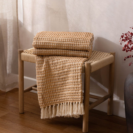 Lorne Plain Cotton Throw in Honey, folded and draped over a woven wooden stool, showing a chunky two-tone waffle weave with fringed edge in a living room setting.