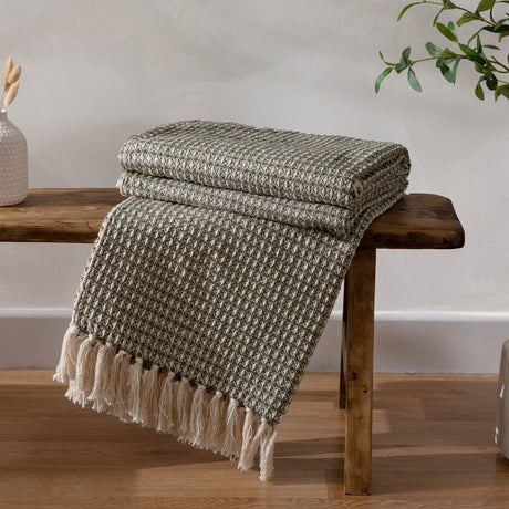 Lorne Plain Cotton Throw in Lichen green, folded and draped with tasselled fringe over a wooden bench in a room with a plant and vase.