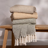 Lorne Plain Cotton Throw in Lichen, folded with waffle-textured green and cream fabric and tasselled fringe, stacked with beige and orange throws on a wooden bench.
