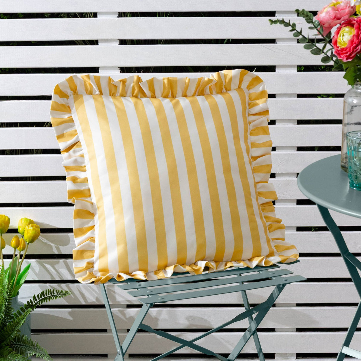 Lottie Striped Polyester Cushion in yellow and white stripes with a gathered ruffle trim, shown on a garden chair against a white slatted fence.