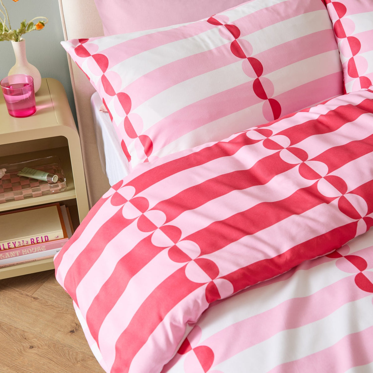 Lottie Reversible Geometric Duvet Cover Set in red and pink, showing striped fabric with vertical half-circle rows on a bed with matching pillowcase in a bedroom setting.