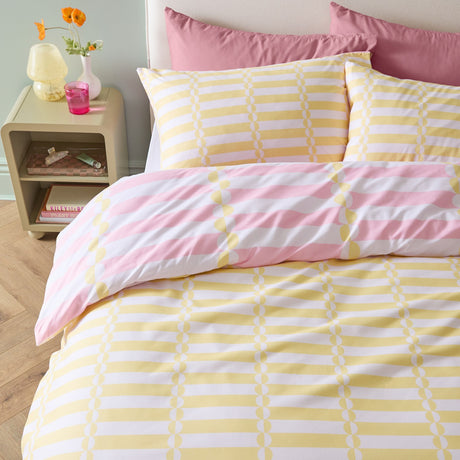 Lottie reversible duvet cover set on a bed, yellow and white striped side with disc pattern shown, pink striped pillowcase reverse visible, in a bedroom setting.