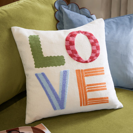 Love Tuft Embroidered Polyester Cushion in cream, 45 x 45cm, with tufted LOVE lettering in green, red-pink check, blue and orange, shown on a green sofa with blue cushions.