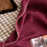 Lowen Wool Plain Polyester Throw in berry red with a dusky pink blanket-stitch edge, draped over a bed with a gingham cover, cushions and a book nearby.