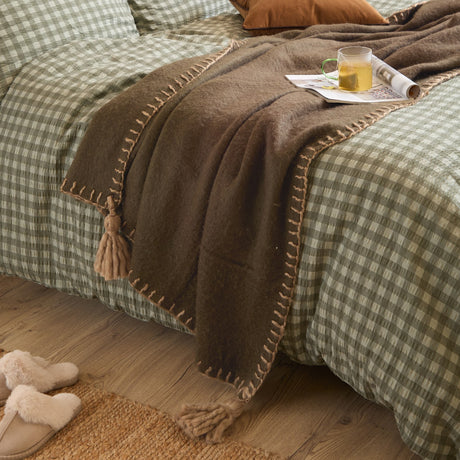 Lowen Wool Plain Throw in chocolate brown draped over a green gingham bed, showing whipstitched edges and tassel corners, styled with a mug and magazine.