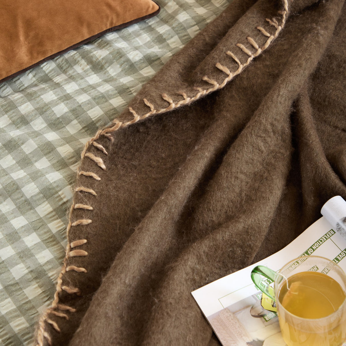 Lowen Wool Plain Throw in chocolate brown with blanket-stitch edging, draped over a green gingham bed near a tan cushion, magazine, and mug of tea.