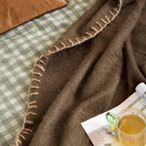 Lowen Wool Plain Throw in chocolate brown with blanket-stitch edging, draped over a green gingham bed near a tan cushion, magazine, and mug of tea.