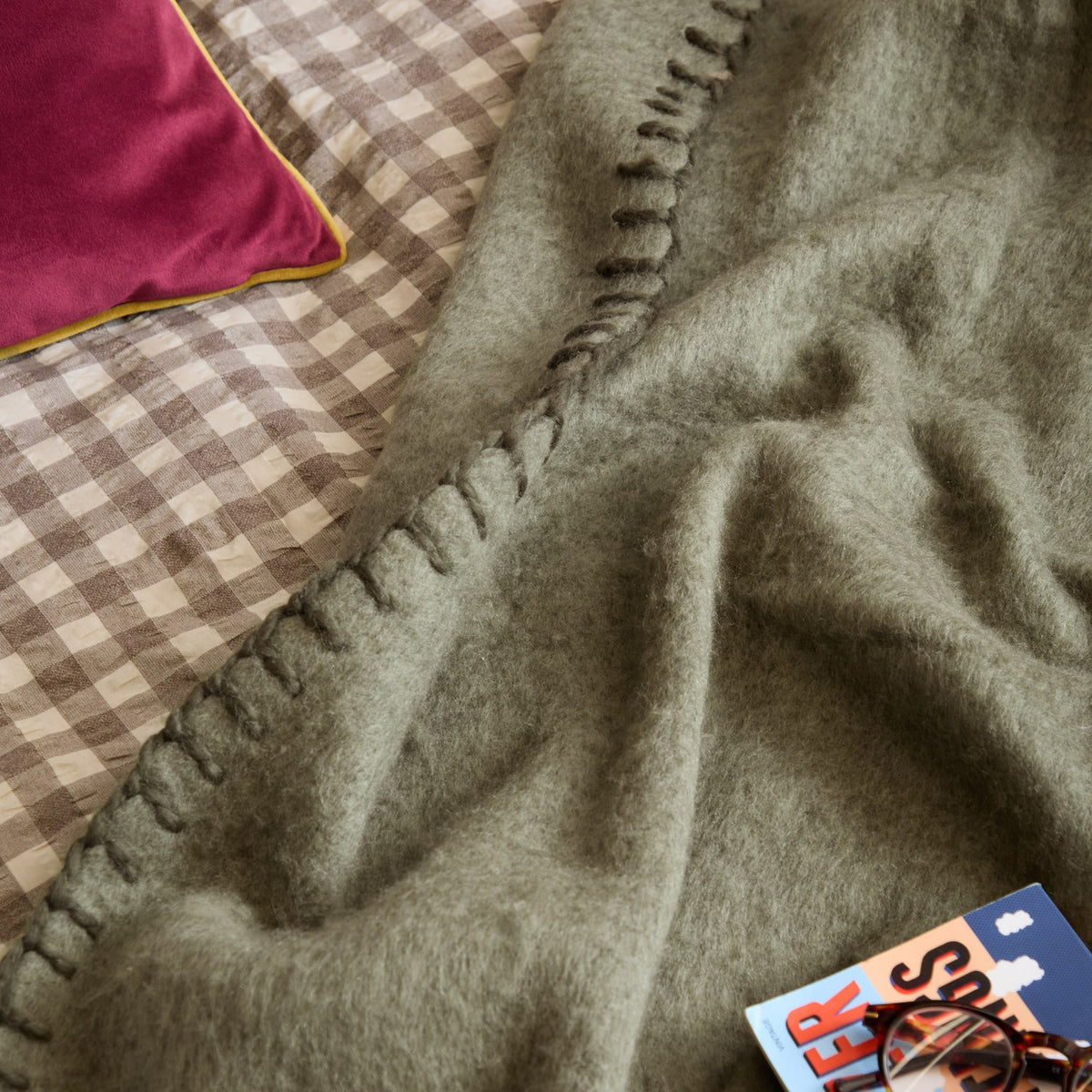 Lowen wool-blend throw in moss green with a whipstitched edge, draped over a bed beside a gingham duvet and pink velvet cushion.