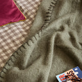 Lowen wool-blend throw in moss green with a whipstitched edge, draped over a bed beside a gingham duvet and pink velvet cushion.