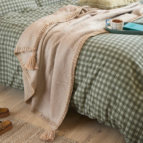 Lowen Wool Plain Polyester Throw in natural beige, draped over a green gingham bed, showing blanket-stitch edging and corner tassels.