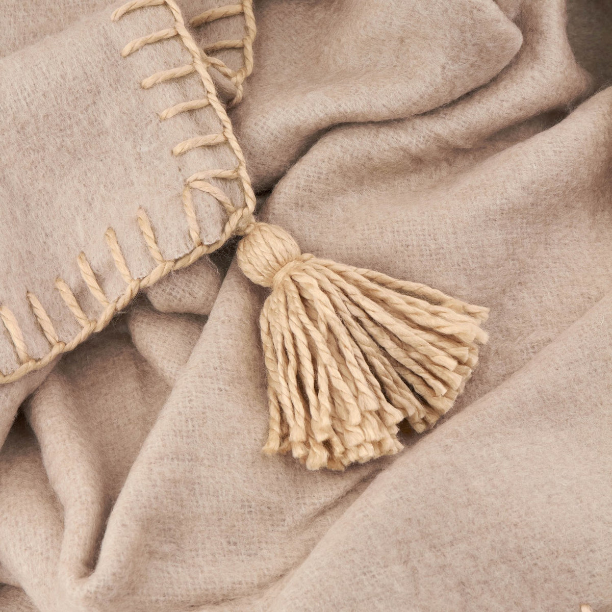 Close-up of the Lowen wool-blend throw in natural beige, showing the hand-worked blanket-stitch edge trim and a jumbo tassel detail on the brushed fabric.