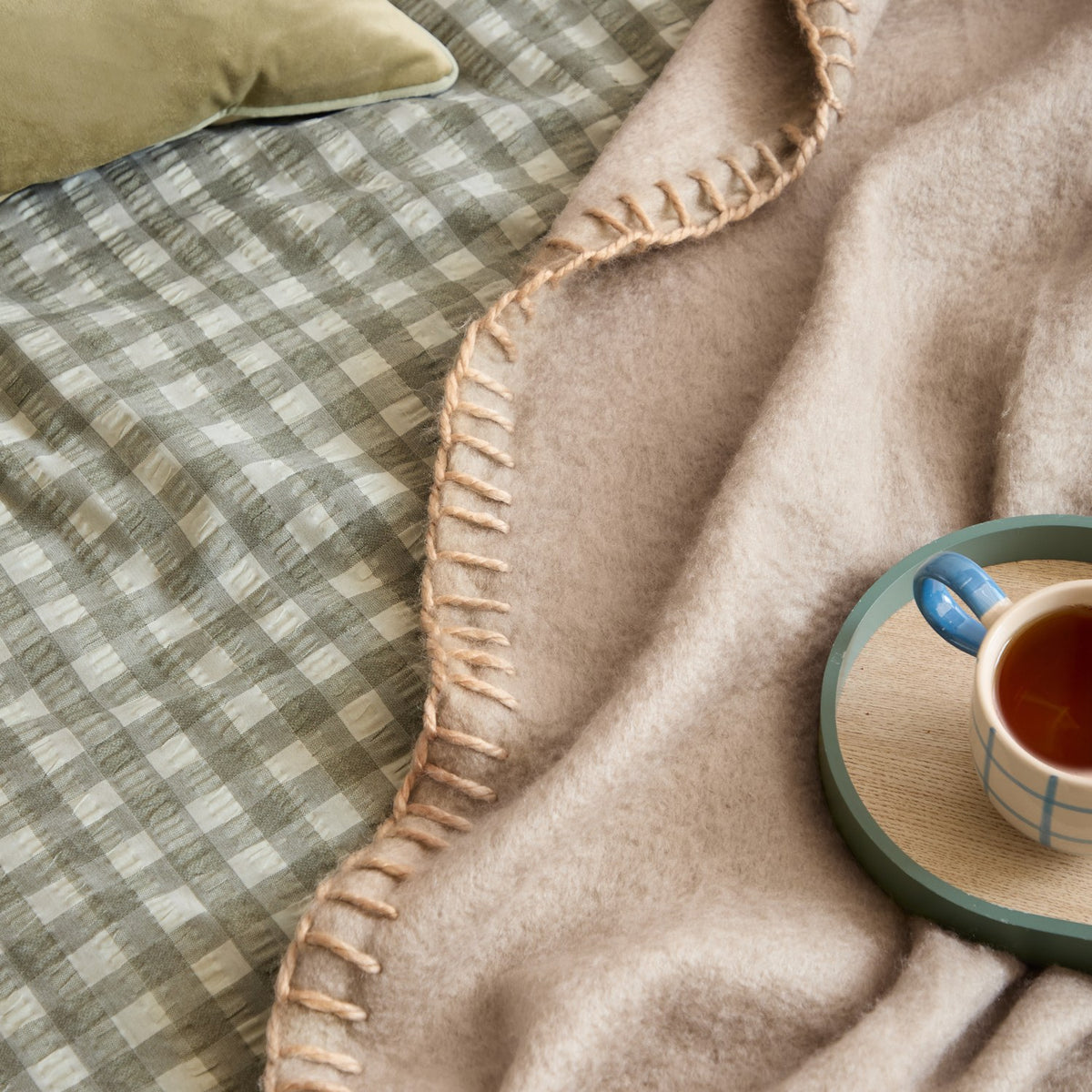 Lowen Wool Plain Polyester Throw in natural beige with hand-worked blanket-stitch edging, draped on a bed beside a green gingham duvet and cup of tea.