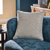 Loxley Velvet Cotton Cushion in Silver, a beige square cushion with metallic speckled foil print, shown on a blue velvet armchair in a living room setting.