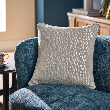 Loxley Velvet Cotton Cushion in Silver, a beige square cushion with metallic speckled foil print, shown on a blue velvet armchair in a living room setting.