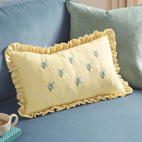 Rectangular yellow and white gingham cotton cushion with frilled trim, embroidered blue flower sprigs, styled on a blue sofa with a teal cushion behind.