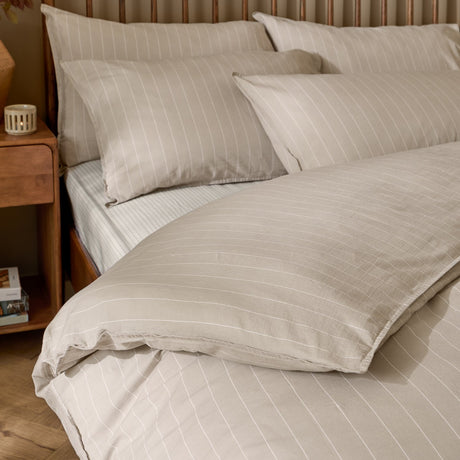 Linear Stripe Cotton Printed Duvet Cover Set in Natural, shown on a bed with matching pillowcases in beige with a fine white pinstripe, styled in a bedroom with a wooden headboard and side table.