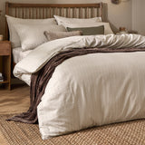 Linear Stripe Cotton Printed Duvet Cover Set in Natural, dressed on a bed with matching pillowcases and a brown throw, in a bedroom with a wooden slatted headboard.
