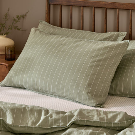 Linear Stripe Printed Cotton Pillowcase Pair in Forest, sage green with narrow white pinstripes, shown styled on a bed with a matching striped duvet against a wooden headboard.