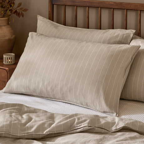 Linear Stripe Printed Cotton Pillowcase Pair in Natural, styled on bedroom pillows with fine off-white lines on a beige cotton ground and a wooden headboard behind.