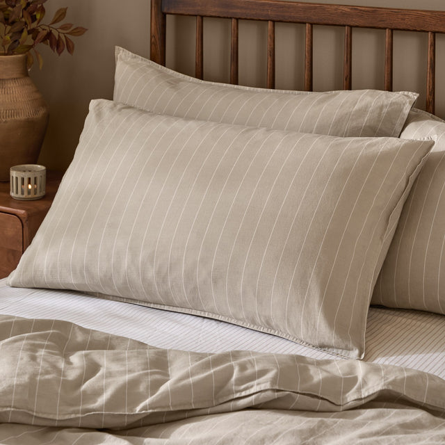 Linear Stripe Printed Cotton Pillowcase Pair in Natural, styled on bedroom pillows with fine off-white lines on a beige cotton ground and a wooden headboard behind.