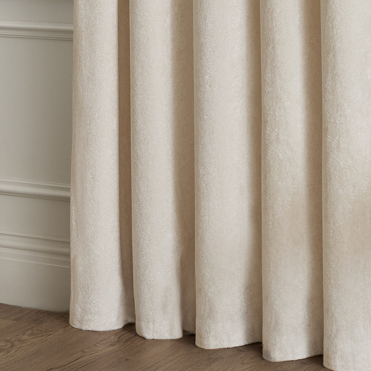 Lustre Velvet eyelet curtains in ivory, close-up of soft pile fabric falling in deep folds, fully lined against a neutral wall and wood floor.