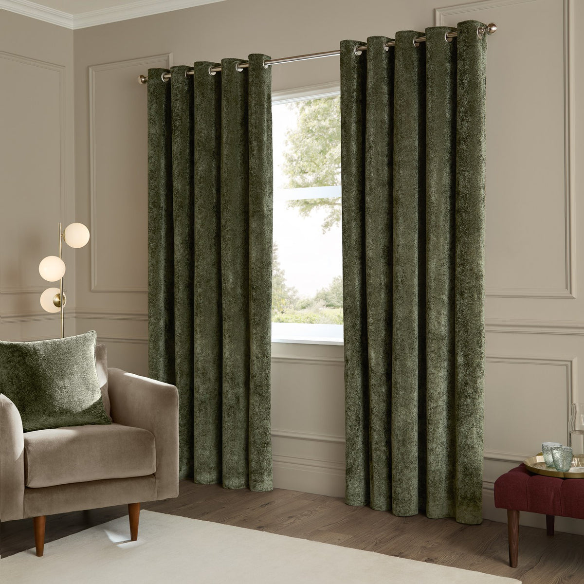 Lustre Velvet Plain Fully Lined Eyelet Curtains in malachite green, shown hung on a chrome pole in a living room beside a beige armchair and window.
