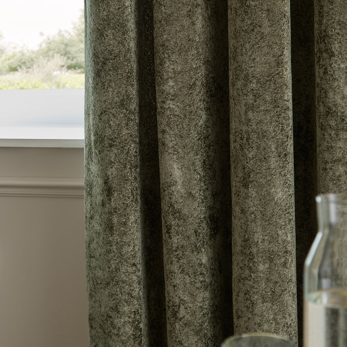 Lustre Velvet eyelet curtains in malachite green, showing the crushed pile texture and heavy folds, hung beside a window with a light interior setting.