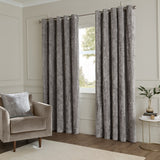 Lustre Velvet Plain Fully Lined Eyelet Curtains in pewter grey, crushed velvet finish, hung on a metal pole in a living room beside a window and armchair.