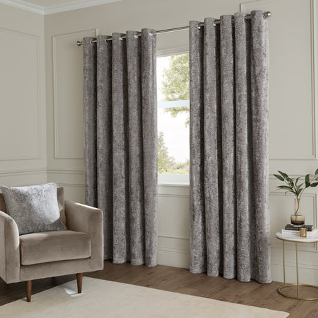 Lustre Velvet Plain Fully Lined Eyelet Curtains in pewter grey, crushed velvet finish, hung on a metal pole in a living room beside a window and armchair.
