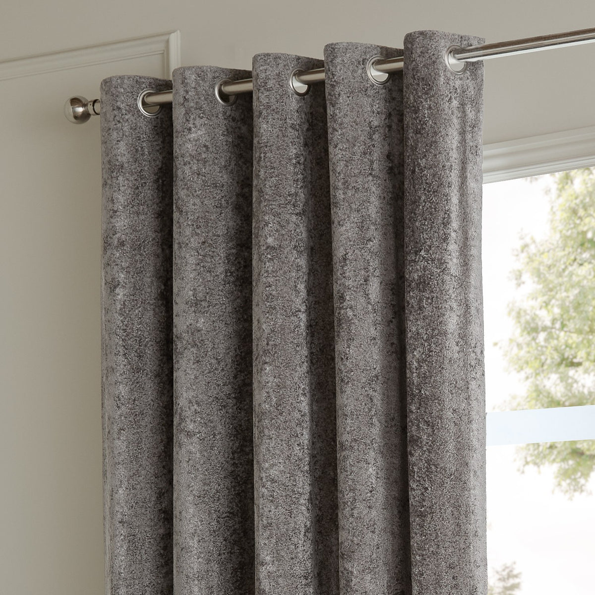 Pewter grey eyelet curtains in crushed velvet fabric, shown threaded on a brushed metal pole beside a window in a living room.