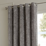 Pewter grey eyelet curtains in crushed velvet fabric, shown threaded on a brushed metal pole beside a window in a living room.