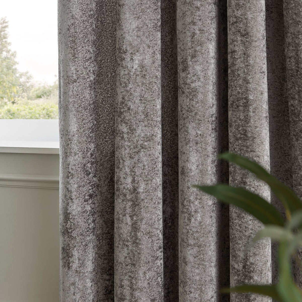 Lustre Velvet Plain Fully Lined Eyelet Curtains in pewter grey, close-up of crushed velvet fabric folds by a window, with greenery visible in the background.