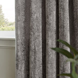 Lustre Velvet Plain Fully Lined Eyelet Curtains in pewter grey, close-up of crushed velvet fabric folds by a window, with greenery visible in the background.