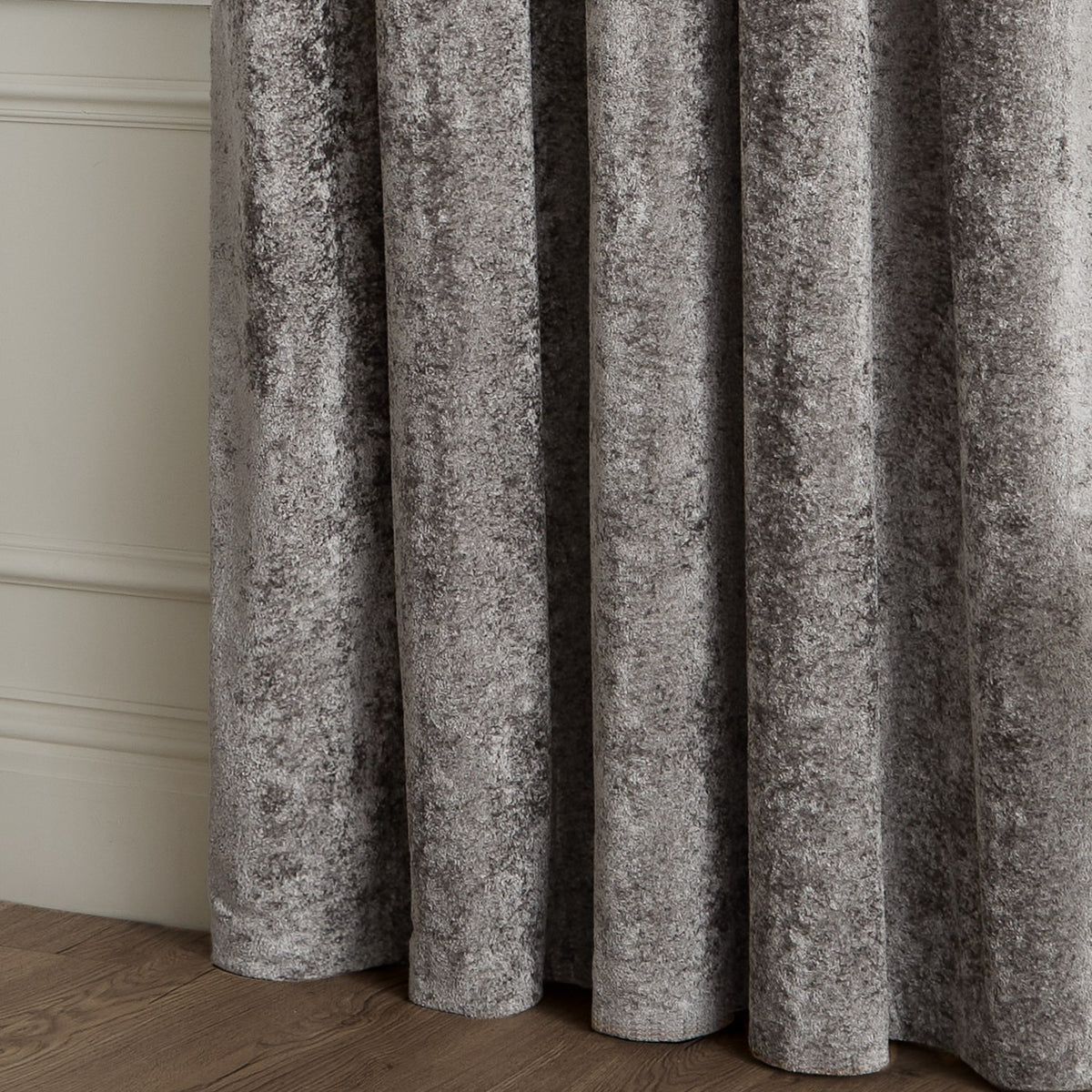 Close-up of Lustre Velvet Plain Fully Lined Eyelet Curtains in pewter grey, showing crushed velvet fabric with soft folds against a panelled wall and wooden floor.