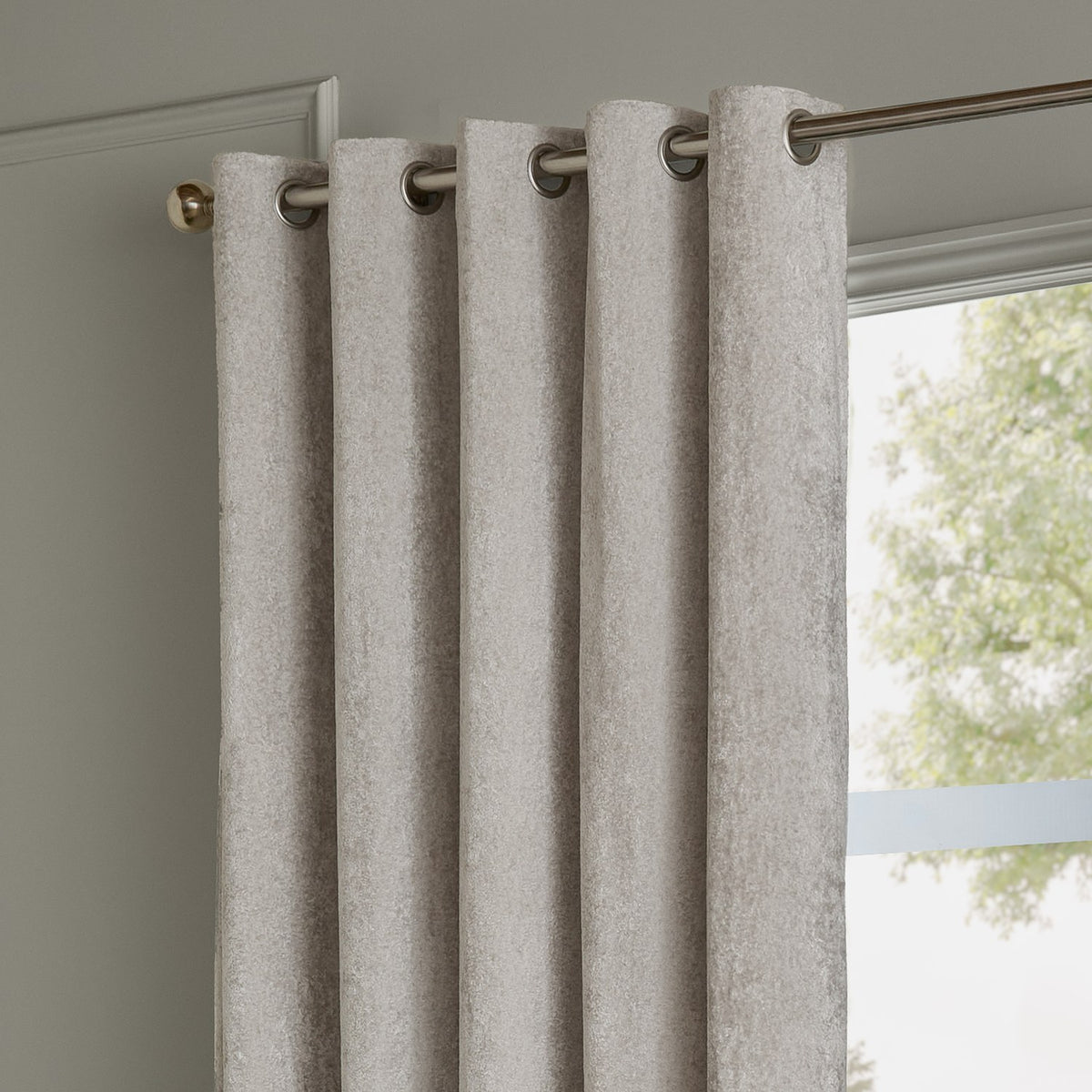 Lustre Velvet eyelet curtain in silver, fully lined with grommet header on a chrome rail, shown hanging beside a window in a light grey room.