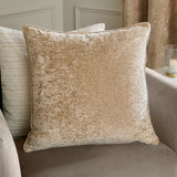Square Lustre Velvet cushion in champagne, showing crushed velvet pile with piped edges, styled on a sofa with a striped cushion and curtain in the background.