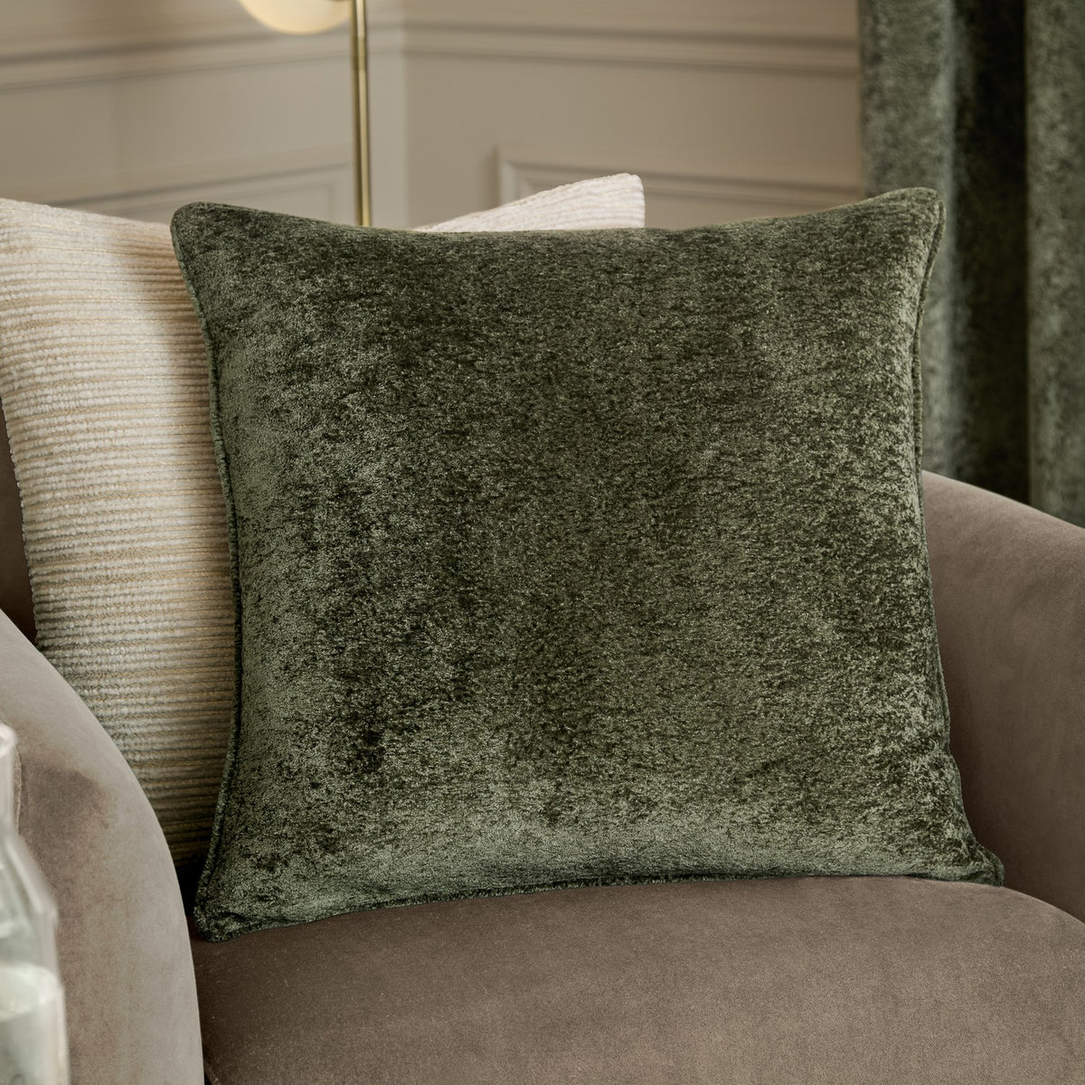 Lustre Velvet Plain Polyester Cushion in Malachite Green, a square crushed-velvet cushion with self-piped edge, shown on a taupe armchair beside a striped cream cushion.