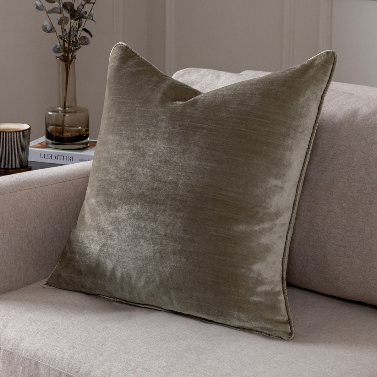 Luxe Velvet Plain Polyester Cushion in mink, a square scatter cushion with piped edges shown on a neutral sofa beside a vase and books.